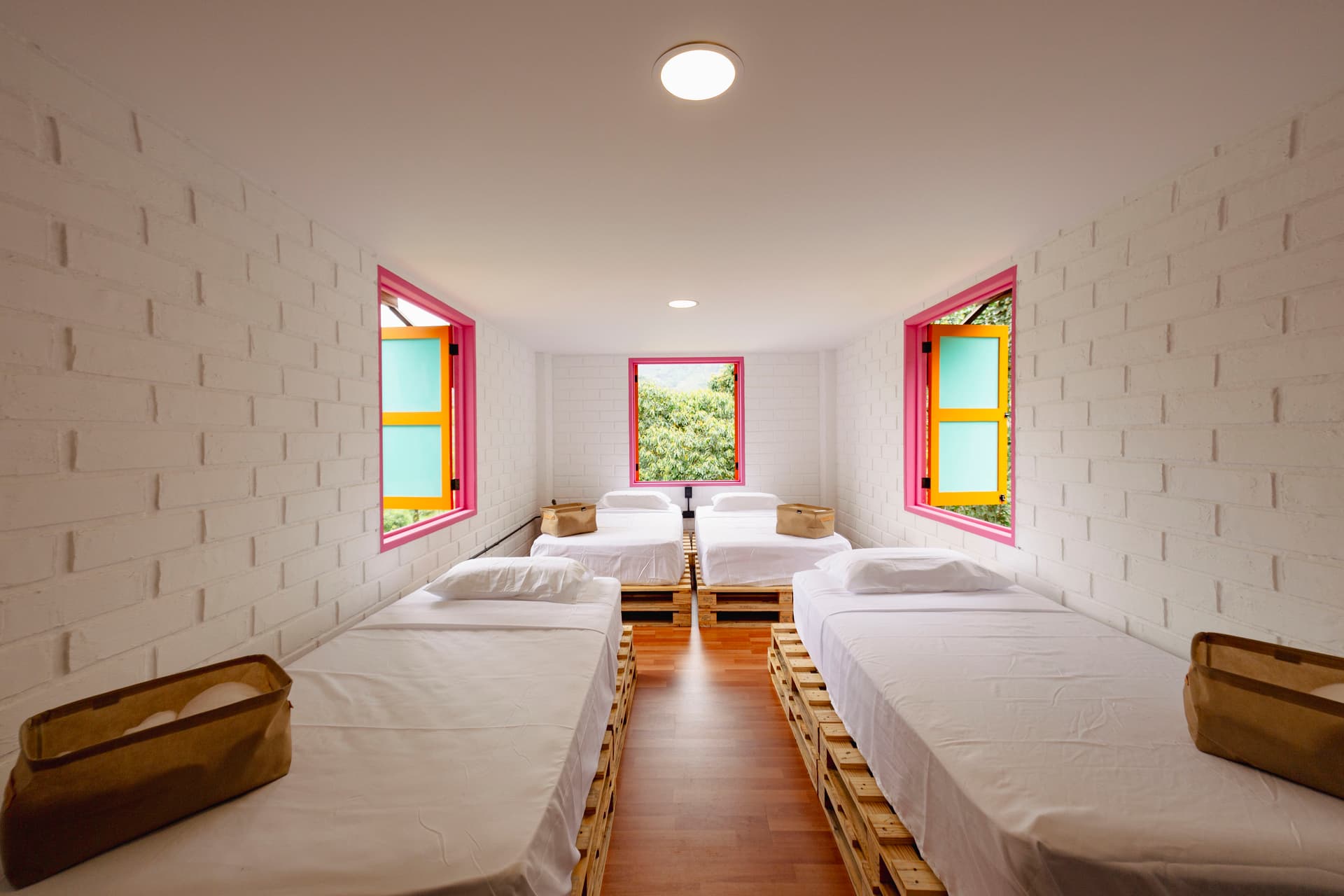 Four single wooden-platform beds beneath three pink-trimmed windows in a white-brick room